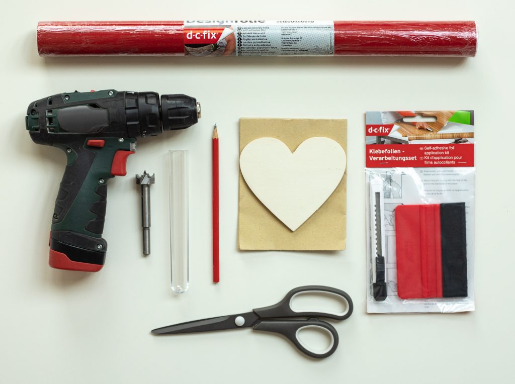 A roll of red adhesive foil, plywood heart, test tube, drill with Forstner bit, sandpaper, pencil, scissors, utility knife and smoothing blade lie on a cutting mat