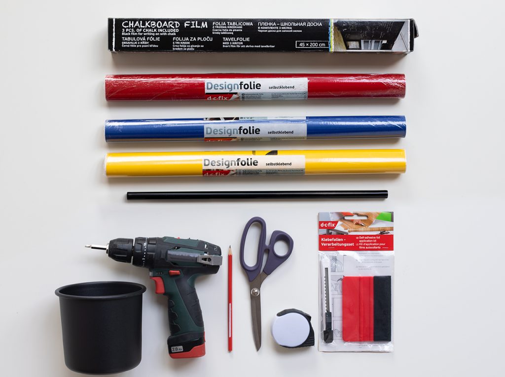 Chalkboard foil, three colorful adhesive foils, chalks, scissors, tape measure, utility knife, scraper, cordless screwdriver, stainless steel rod and three round containers on a top.
