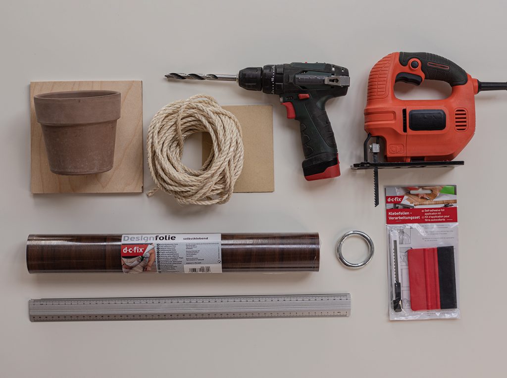 A roll of Walnut adhesive foil, 3 equal square wooden panels, 3 pots and plants, cord, metal ring, drill, jigsaw, sandpaper, ruler, utility knife and a scraper on a worktop.