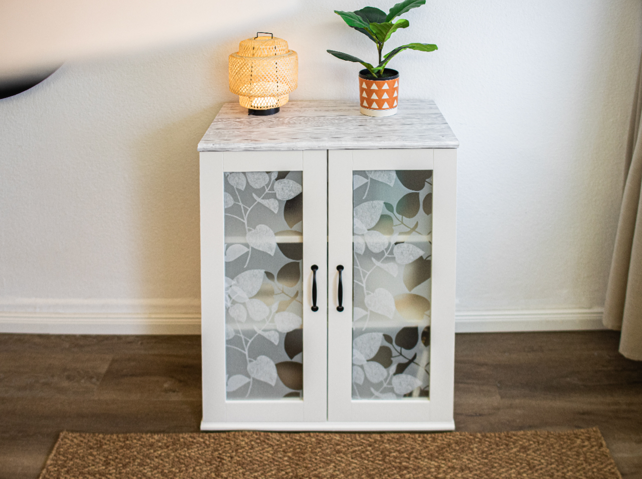 White cabinet with storage shelf in vintage white wood look. The two doors of the cabinet feature glass inserts with semi-transparent window foil bearing a leaf pattern.